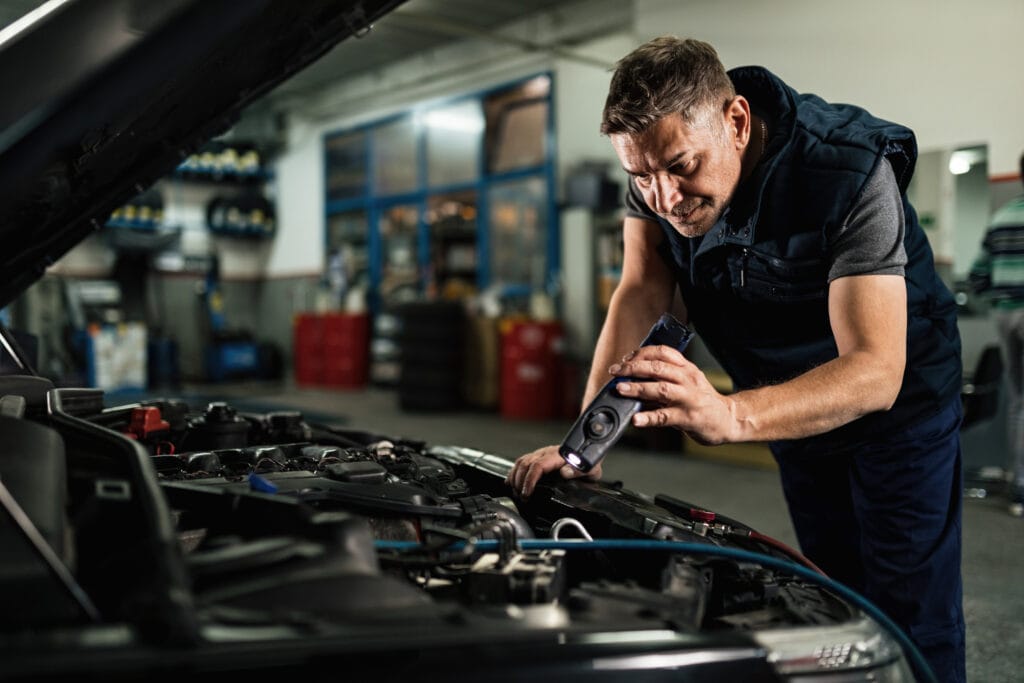 Mid adult mechanic examining car engine while using a lamp in auto repair shop.